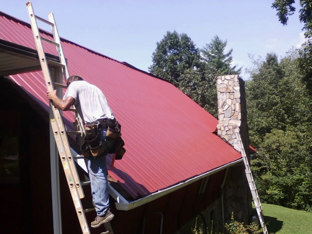 Roofer on a ladder beside a red metal roof and stone chimney — Soffit & Fascia Repair by Roofing Brothers in Blackville